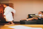 Person im Bademantel geht an einem auf der Couch Lesenden vorbei