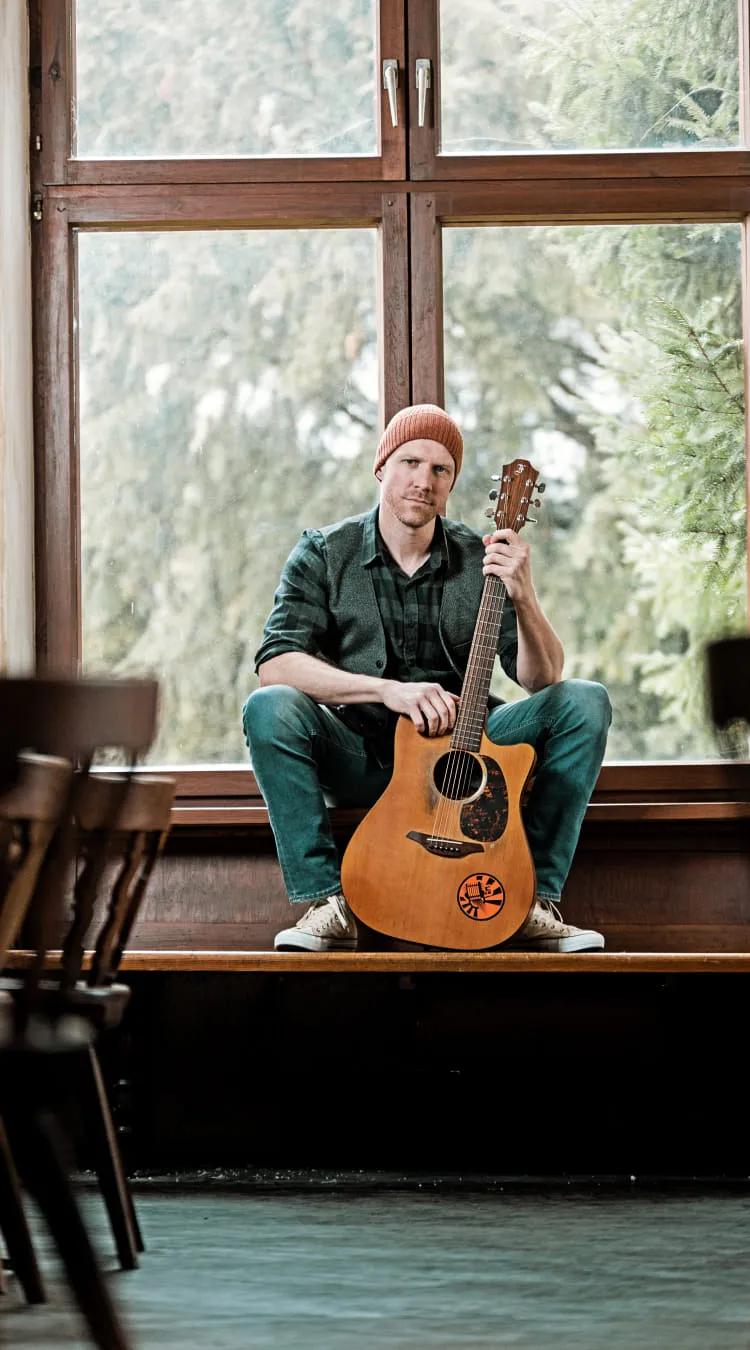 Mann mit Gitarre sitzt auf Fensterbank in einem Raum.