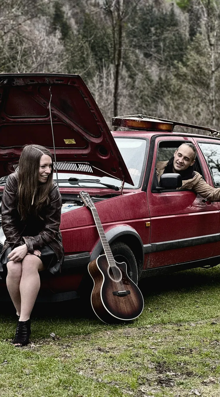 Zwei Personen mit Gitarre neben einem roten Auto.