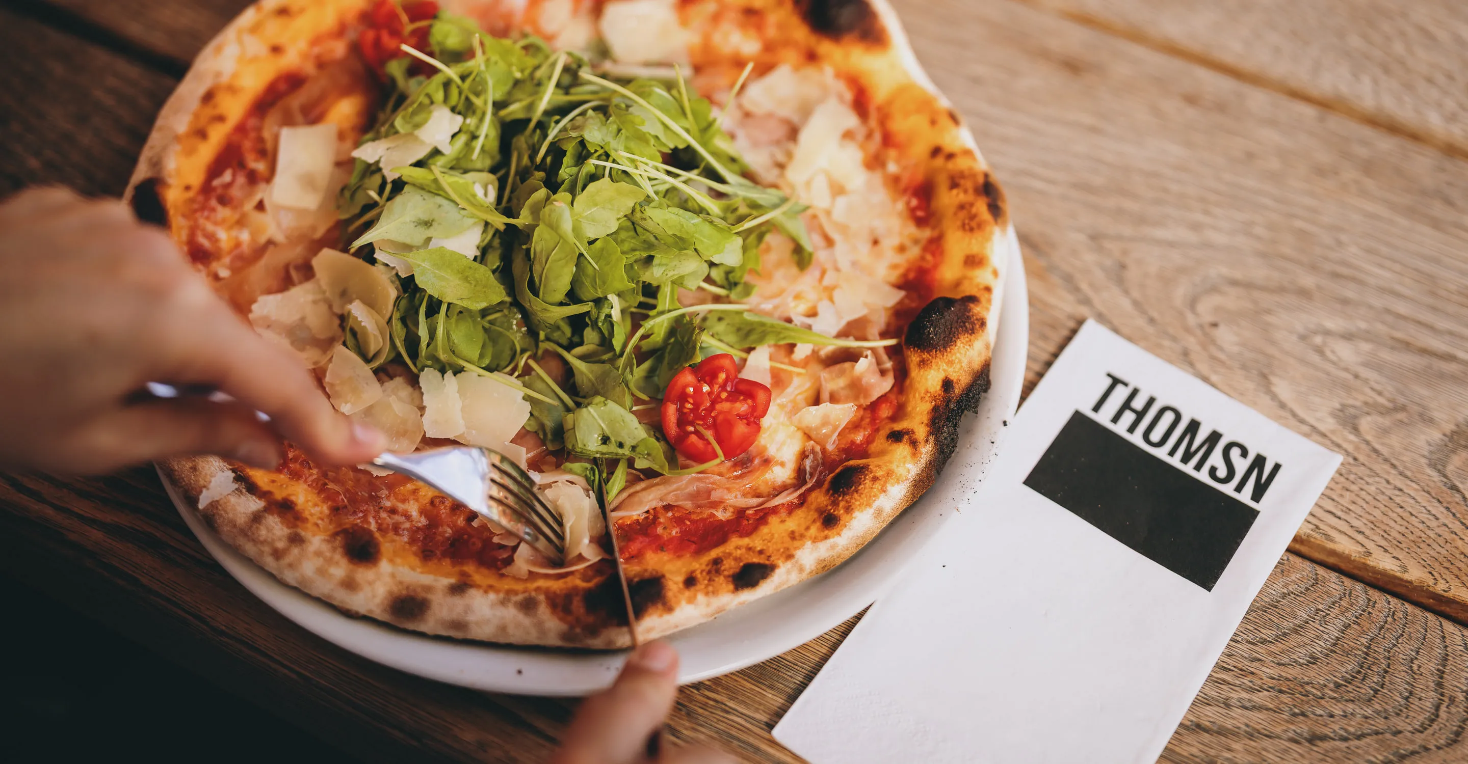 Eine Person schneidet eine Pizza mit Salat auf einem Tisch bei THOMSN.