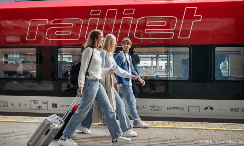 Three travelers walk by the train platform with a railjet.